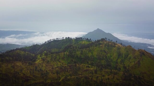 Lush green volcanic ridges rise above a low cloud layer with a distant peak fading into the hazy sky in Bali, Indonesia.