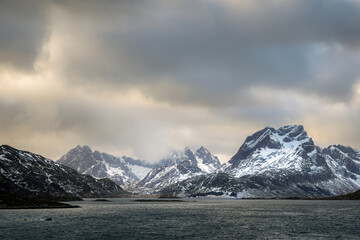 Snow-covered jagged peaks across dark Arctic water beneath heavy cloud, Norway © dachakrapan