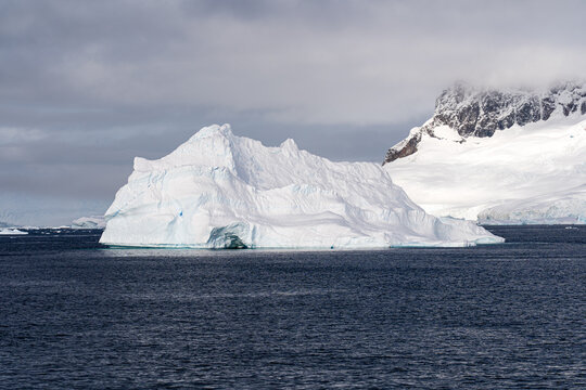 Large iceberg floats in dark water with grey skies above. Antarctic expedition. Eckener Point. Zodiac landing