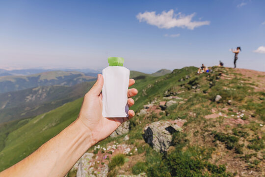 Hand holding a white energy drink pouch against a mountain landscape with hikers under a clear sky