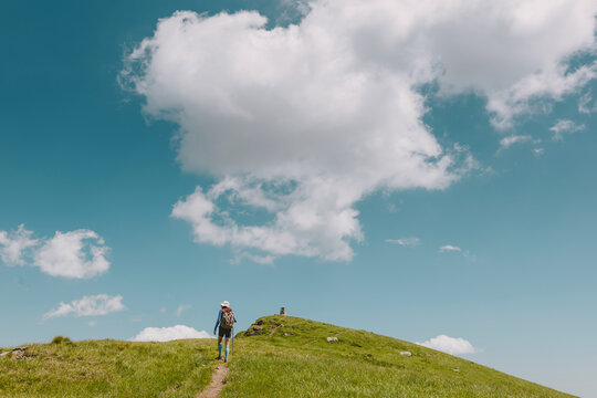 Person hiking with a backpack and trekking poles on a grassy hill, exploring nature under a blue sky with clouds