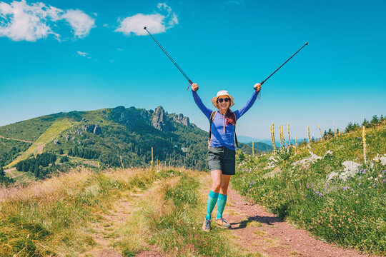 Happy woman hiker with trekking poles raising arms in victory gesture, standing on a mountain trail under a clear blue sky
