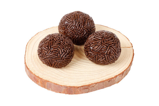 Three traditional Brazilian chocolate brigadeiros coated in sprinkles, on a round wooden base, isolated against a white background.