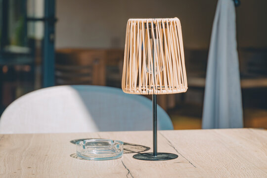 Rattan lamp casts warm morning sunlight and shadows across a wooden table with an ashtray on an empty outdoor cafe terrace