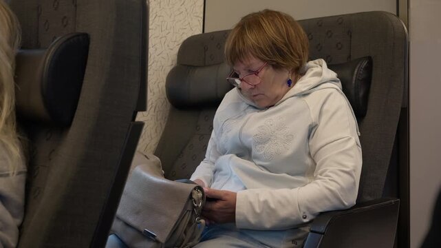 A tired elderly woman wearing eyeglasses dozes off on a passenger train seat. She rests with a smartphone and handbag during a daily commute. A mature senior passenger naps on public transit.