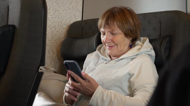 An elderly woman checks her smartphone while traveling by train. A mature passenger manages online services, tickets, and bookings on a trip. She enjoys independent modern transportation.