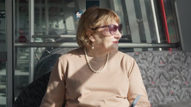 An elderly woman in sunglasses sits by a window inside a passenger train. A mature senior passenger enjoys a calm commuter rail journey. She looks outside while traveling across the city.