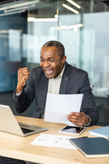 Obraz premium Happy mature executive celebrating success with a fist pump, looking at documents and a laptop, enjoying a triumphant moment in a modern corporate office environment
