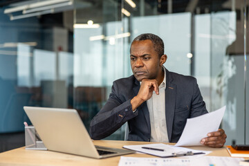 Obraz premium Experienced entrepreneur focusing on analyzing business documents while using a laptop at an executive desk, planning financial strategies and making important decisions