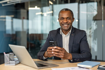 Obraz premium Mature professional manager engaging with technology in a modern corporate office, confidently communicating and enjoying a successful workday while using a mobile device at his desk