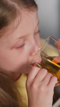 Close up of young girl drinking from glass with golden liquid. Girl holds transparent cup near her mouth focusing on eating activity. Soft lighting highlights details in close up shots