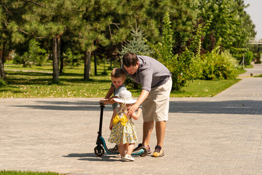 Father supports his young son on a kick scooter while gently guiding his toddler daughter standing beside them during a sunny family outing.