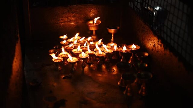 Cluster of brass butter lamps burns in a darkened shrine niche, warm flames reflecting on stone, Kathmandu, Nepal