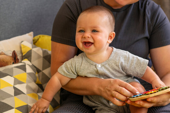 Happy infant smiles while sitting on a caregiver's lap as the woman holds and demonstrates a colorful wooden gear toy during interactive learning and play at home.