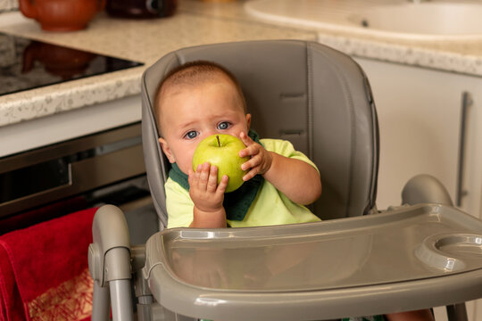 Baby Boy Holds Green Apple In Kitchen High Chair