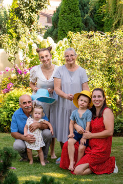 Grandparents, two adult women, and two young children pose together for a warm multigenerational family portrait in a flowering summer garden.  