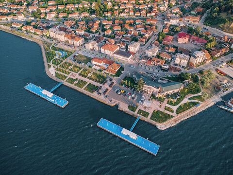 Aerial view of a peaceful riverside town with terracotta rooftops, waterfront promenade, docking piers, and a barge moored along the Danube river. Travel concept.