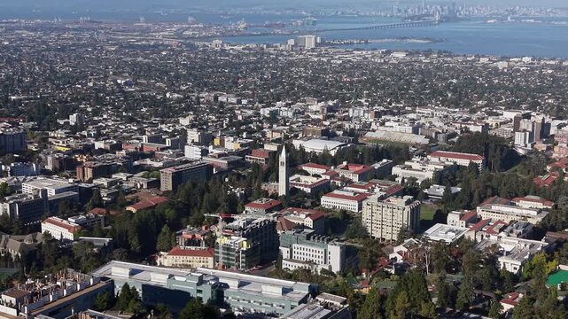 Aerial drone glides over UC Berkeley&rsquo;s campus, showcasing iconic Sather Tower, lush trees, sprawling cityscape, and sparkling San Francisco Bay under vibrant sunlight.