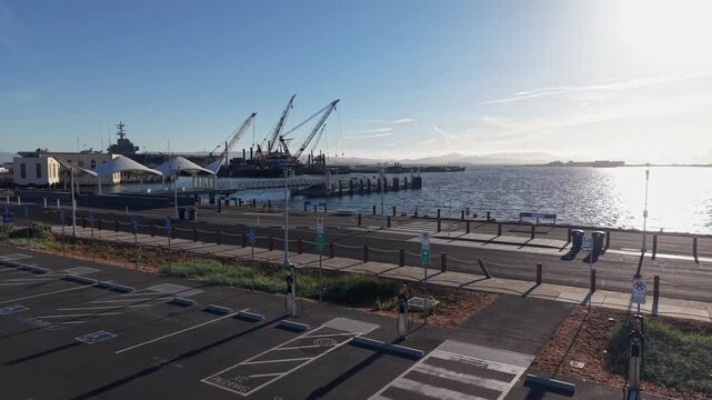 Aerial view ascending over an empty parking lot and ferry terminal at Alameda Point, revealing an EV Charging station, the waters of San Francisco Bay and the USS Hornet Museum on a bright, sunny day
