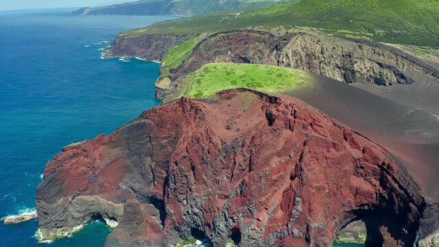 A deep red cliff pierced by sea arches meets a slope of black ash at Capelinhos on Faial Island above deep blue water.