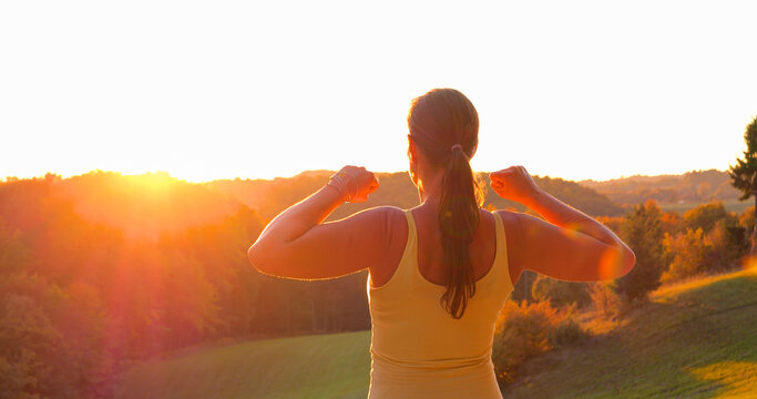 CLOSE UP, LENS FLARE: Back view of young woman raising clenched fists in victorious pose above colourful autumn hills. Powerful moment of achievement and celebration after an active day in nature.