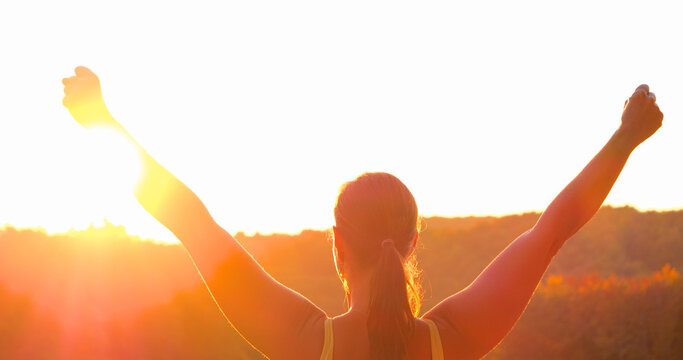 CLOSE UP, LENS FLARE: Young woman lifts her arms in celebration while overlooking glowing autumn countryside. Pristine moment of victory, strength and accomplishment after an active day in nature.
