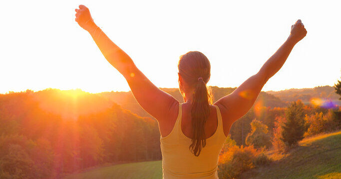 CLOSE UP, LENS FLARE: Woman on a scenic hill with both arms lifted in victorious pose. Warm golden sunlight and autumn colors spill over forested valley as she celebrates her sporting achievement.