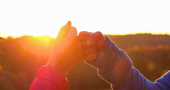 CLOSE UP, LENS FLARE: Two adult people greet each other with a fist bump in warm golden sunlight. Moment of bonding and celebration with forested hills in the background and glowing morning atmosphere