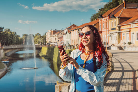 Young woman with red hair and sunglasses happily using her smartphone for navigation, enjoying a sunny city trip by a canal in Knjazevac, Serbia