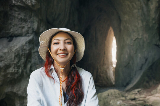 Woman wearing a bucket hat and a white shirt, smiling while standing in a dimly lit cave with a light source visible