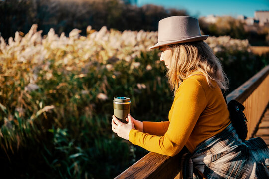 Thoughtful woman with thermo mug on wooden bridge &mdash; eco friendly habits and sustainable lifestyle concept. Relaxed female traveler holding warm drink outdoors. Autumn coffee break, slow living.