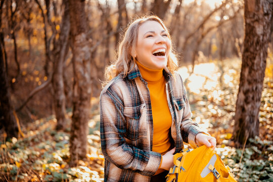 Cheerful woman with yellow backpack resting in autumn forest - active recreation choice. Joyful female hiker enjoying bright sunny weather outdoor. Mental well-being and digital detox concept.