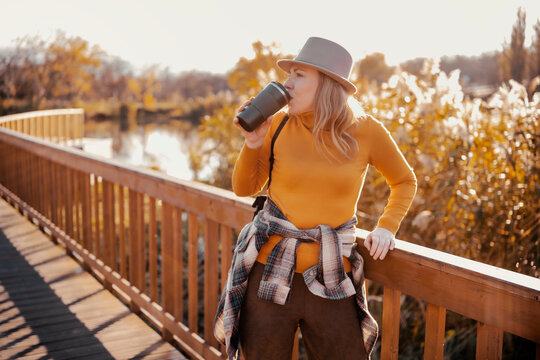 Smiling woman drinking coffee from thermo mug on wooden park bridge &mdash; autumn outdoor wellness and cozy lifestyle concept. Female traveler sipping warm tea in nature, slow living, solo trip.