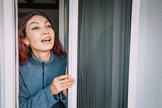 Smiling woman looking through a mosquito screen door onto a sunny balcony, enjoying fresh air and protection from insects during summer