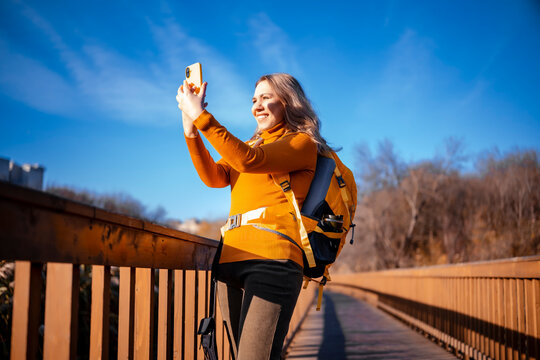 Tourist woman making selfie picture in autumn forest on wooden path - travel blogging. Female hiker recording video content for social media. Modern technology and networking concept.
