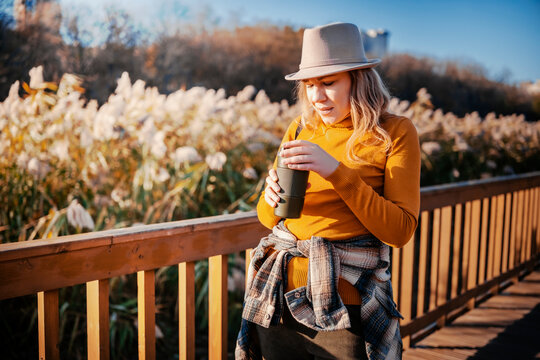 Autumn traveler woman with eco cup on wooden bridge - sustainable lifestyle. Female tourist holding warm drink in nature. Slow living and mindful consumption trend during weekend getaway.