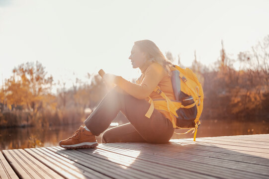 Relaxed woman with mobile phone sitting by autumn lake - peaceful outdoor recreation. Female traveler messaging online in sunny nature park. Work life balance and mental health concept.
