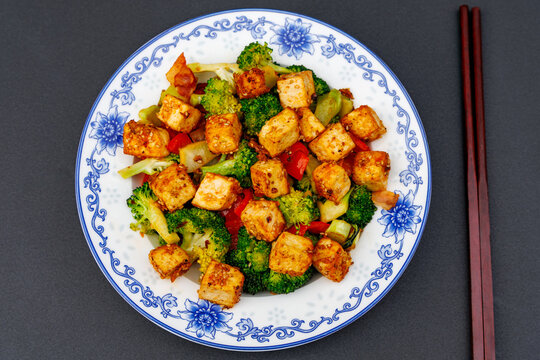 Top down flat lay view of fried crispy tofu stir fry with broccoli on blue porcelain plate with chopsticks, Asian vegan healthy food concept