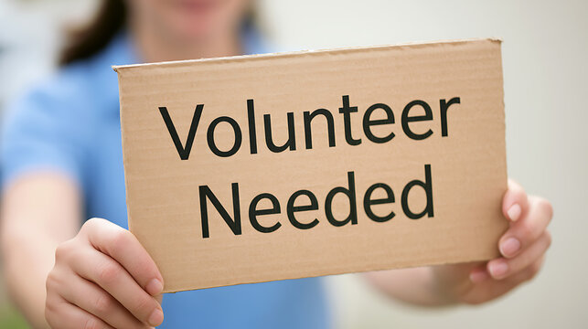 A close up view of a person holding a cardboard sign with black text that reads volunteer needed for community service and charity work
