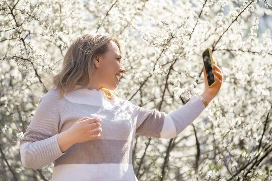 Woman takes smartphone selfie, standing under white spring blooming trees. Springtime outdoor mobile communication lifestyle concept.