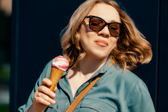Attractive woman in sunglasses eating ice cream cone outdoors &mdash; summer street food and urban lifestyle concept. Joyful female tourist enjoying sweet dessert, city vacation and positive emotions.