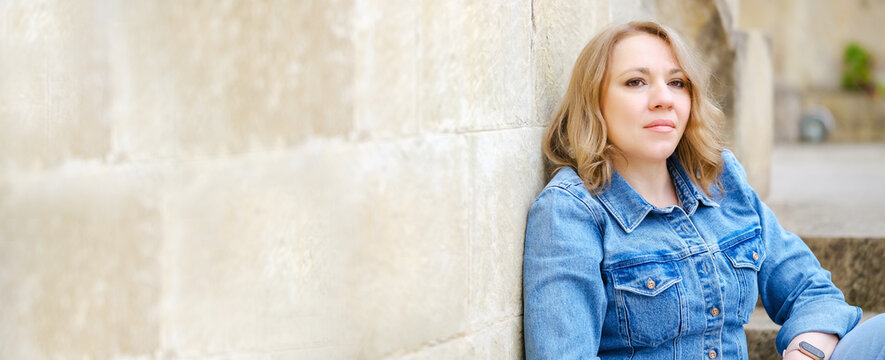 Calm woman in denim jacket poses near stone wall. Beautiful blonde woman looking at camera outdoors.