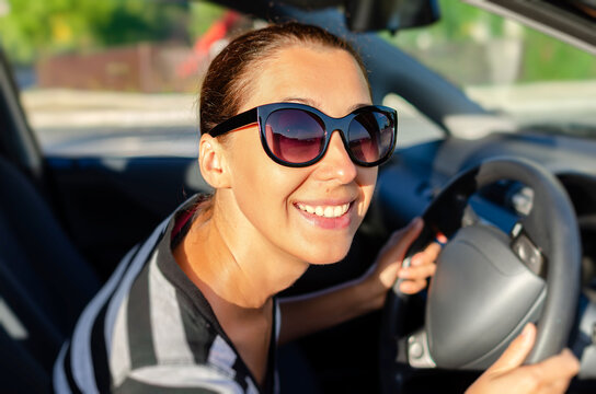 A cheerful woman wearing dark sunglasses smiles while holding the steering wheel of her car on a sunny day.