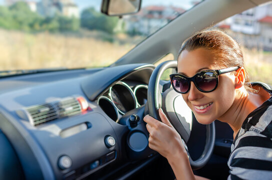 A smiling young woman wearing sunglasses holds the steering wheel while sitting inside her car on a sunny day.