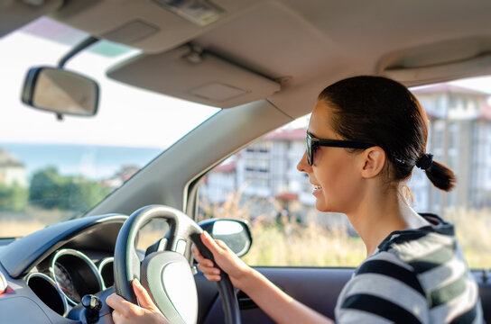 A young woman wearing sunglasses holds the steering wheel while driving a car along a road on a sunny day.