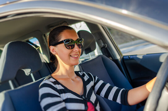 A smiling young woman wearing sunglasses drives a car on a sunny day.