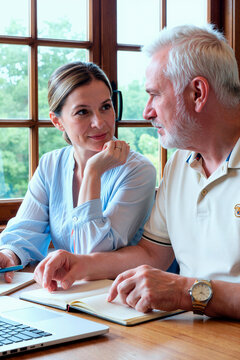 Middle aged Caucasian woman and senior Caucasian man discussing business while working with laptop and notebooks. Image suitable for retirement planning or financial advisory services, vertical