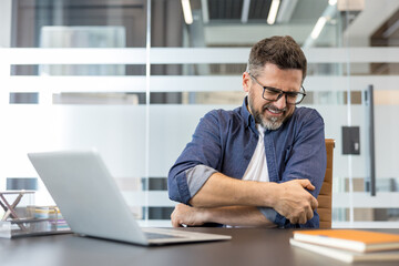 Obraz premium Male office worker experiencing acute pain and discomfort in his elbow while sitting at a desk with a laptop, indicating a health problem or injury from repetitive strain