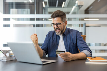 Obraz premium Man in glasses celebrating a successful online payment on his laptop at a modern office desk, smiling while holding a credit card after completing an e-commerce purchase