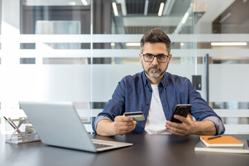 Obraz premium Man in office setting holding a credit card and smartphone, engaging in secure online shopping and mobile banking transactions, using digital financial technology